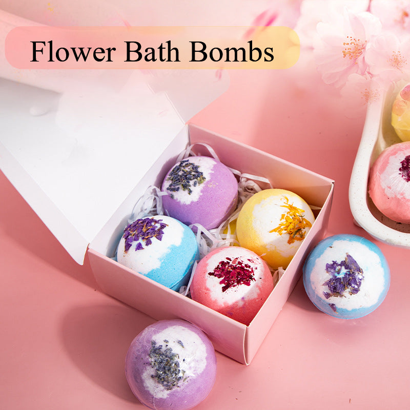 Vier Blüten-Badebomben in Rosa, Gelb, Blau und Violett in einer offenen rosa Geschenkbox; weitere Badebomben sind daneben dekoriert.